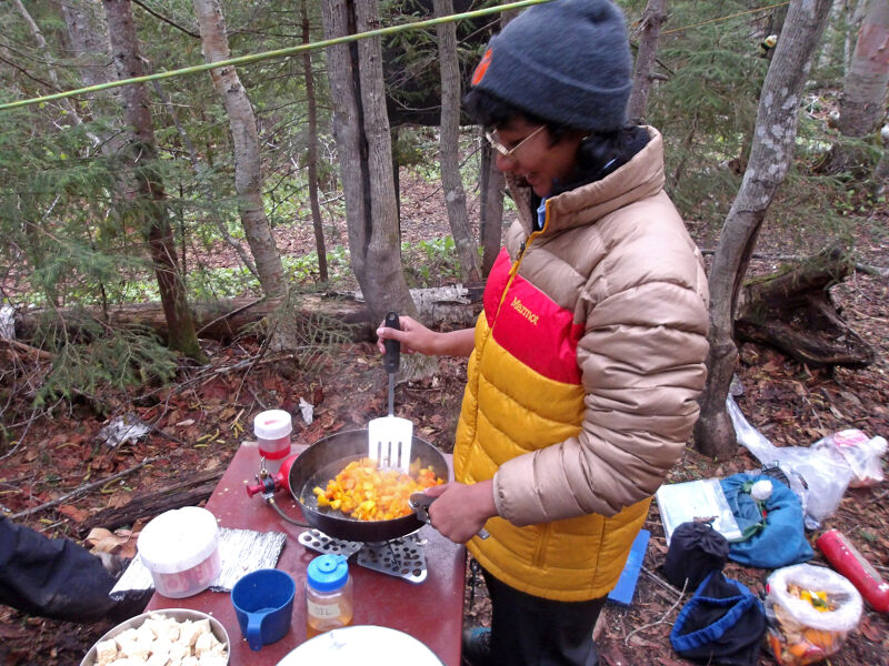 A person wearing a beanie, glasses, and a puffy jacket is cooking food in a frying pan outdoors. The person is standing at a red table with various containers and cooking supplies. The surrounding environment appears to be a wooded area, suggesting they are camping or cooking in nature.